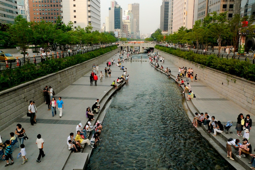 A man made stream weaves through an urban setting with people sitting around the banks