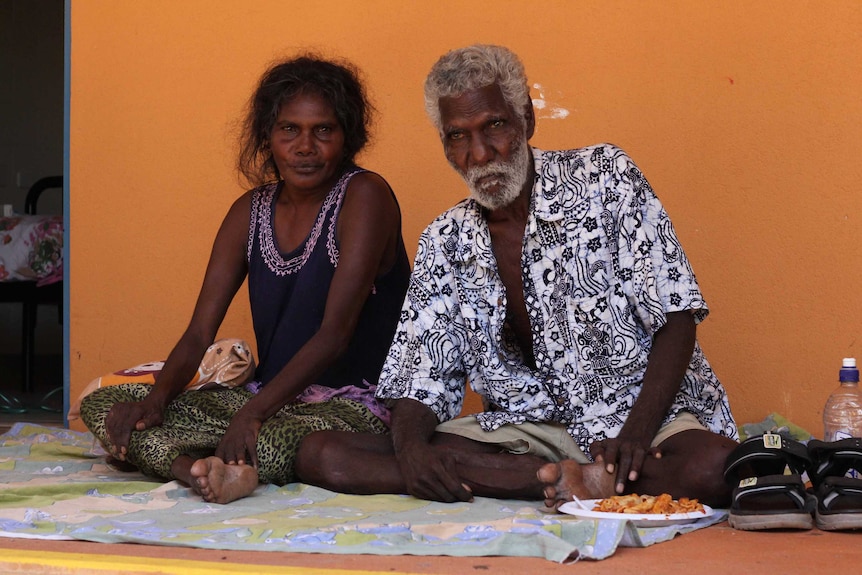 An elderly man and his younger wife sit on the ground on a blanket.