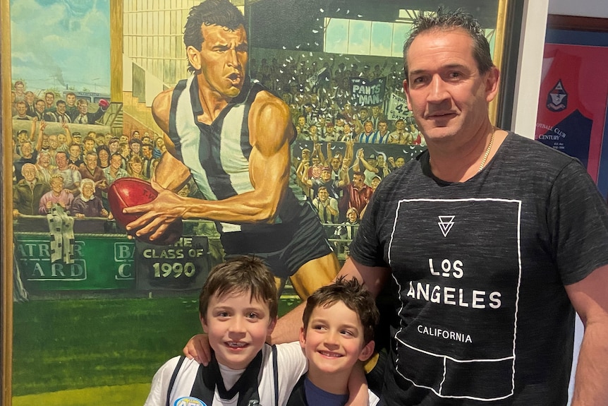 Sean Millane and his two sons stand alongside a painting of his brother Darren playing for Collingwood.