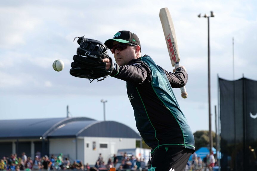 A cricket coach holds a bat in hand, getting ready to hit a ball to fielders in a drill.