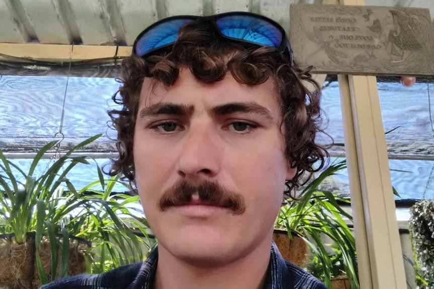 A young man with sunglasses on top of curly brown hair and a mustache looks seriously into the camera. 