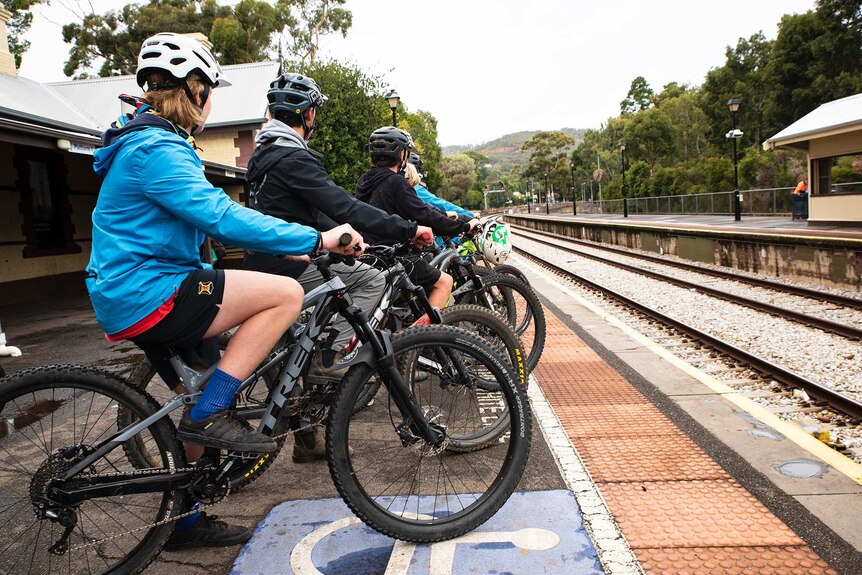 A group of teenagers sit on their bikes on a railway station platform.