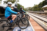 A group of teenagers sit on their bikes on a railway station platform.