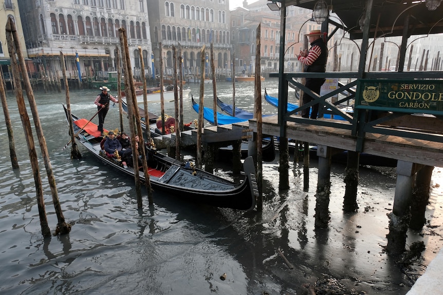 A black gondola sits on a muddy surface at a dock on Venice's Grand Canal, as traditionally dressed gondoliers look on.