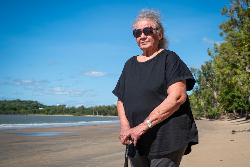 A woman wearing black and dark sunglasses stands on a sandy beach