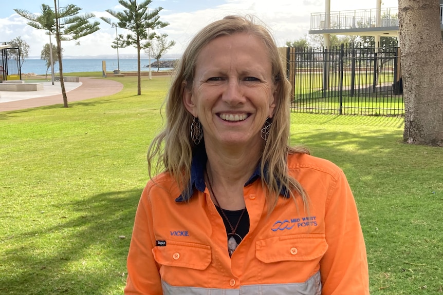 Woman with long hair and fluoro orange work shirt, smiling on foreshore with water, grass and trees behind.
