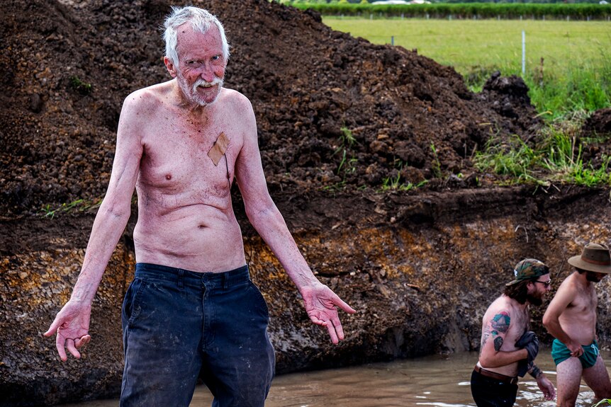 An elderly man stands shirtless near piles of dirt.