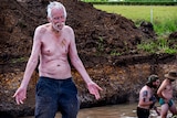 An elderly man stands shirtless near piles of dirt.