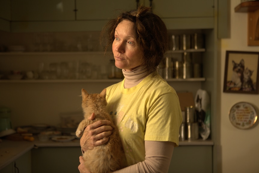 Middle-aged woman with dark hair pinned loosely in plaits and wearing a shirt over a skivvy holds a cat and stares vacantly.