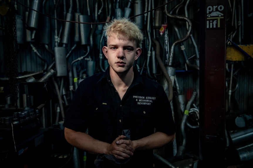Teenage boy in car mechanic uniform stands in dim-lit garage. 