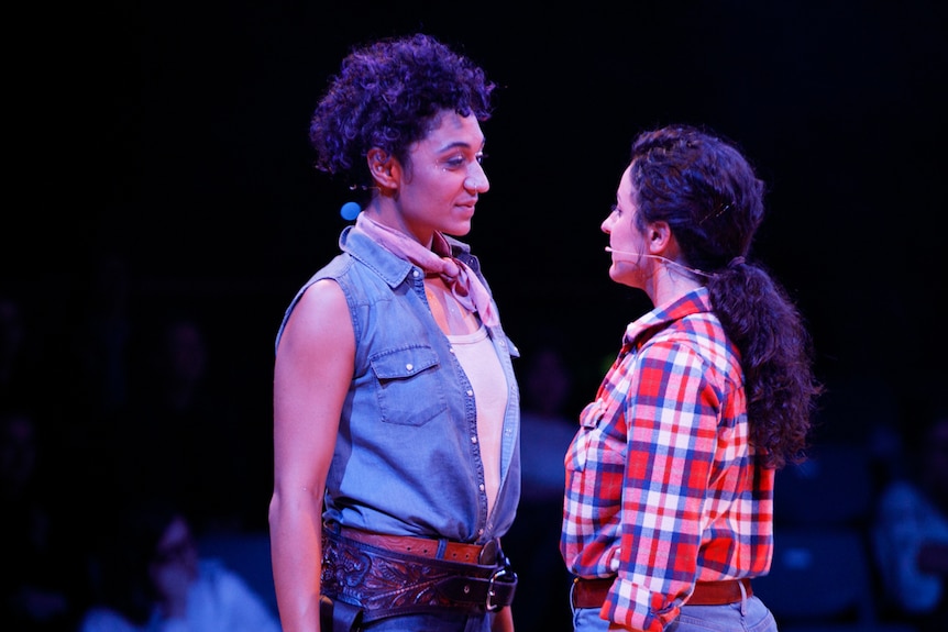 Two women in cowboy-style clothes on a pink-lit stage, wearing face mics, standing close and staring into each other's eyes.