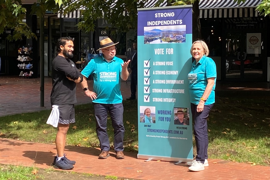 A man and a woman in matching shirts stand talking to another man in front of a sign that reads "Strong Independents".