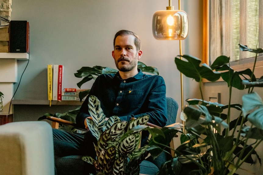 A man wearing a dark shirt sits among house plants next to a window.