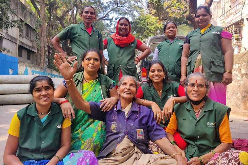 A group of Indian women in green worker unions pose together in the street smiling at the camera. 