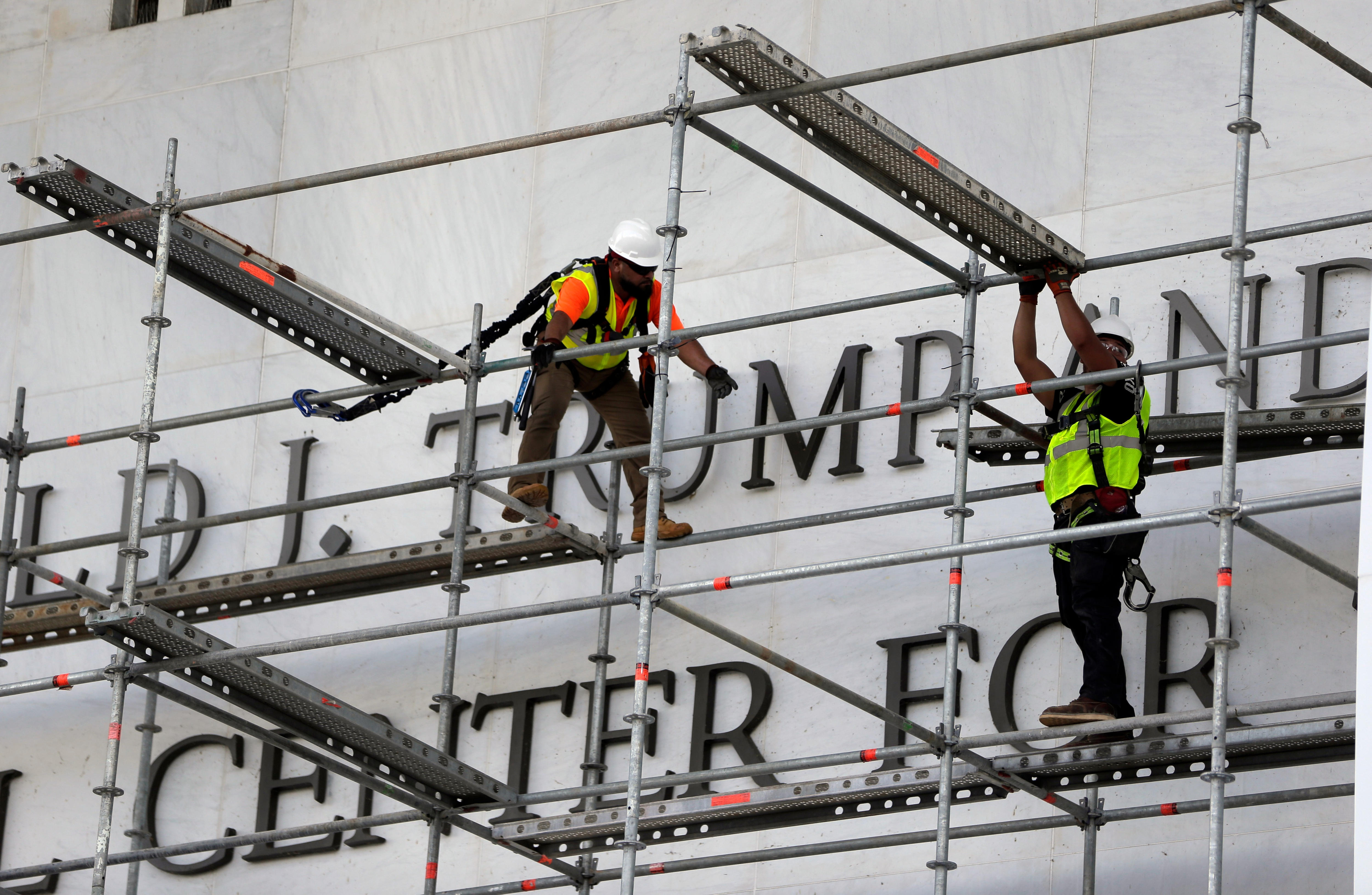 Two men in yellow hi-vis clothing and white hard hats stand on scaffolding in front of a large metal TRUMP sign.