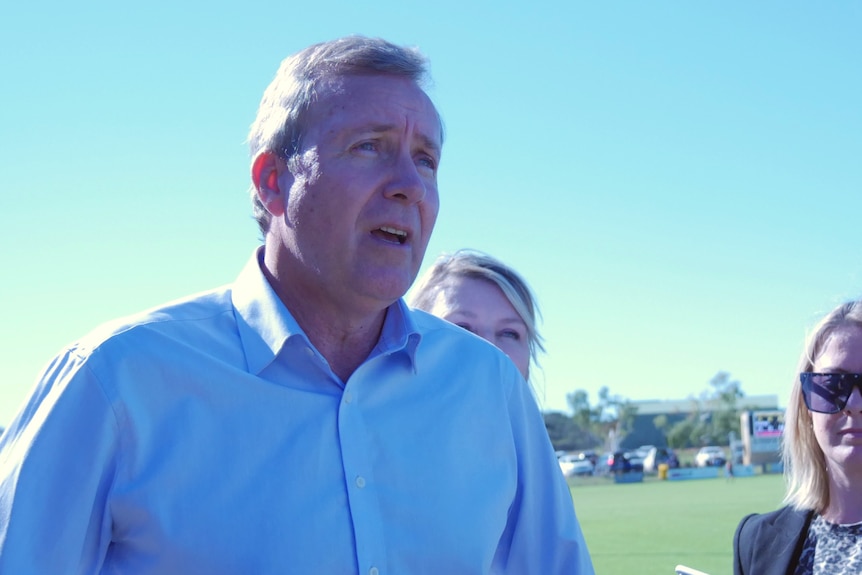 A man wearing a blue shirt is speaking, with a football field in the background.