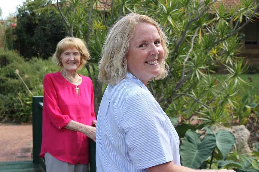Margaret Aiston in background smiling with Janice Millington in the foreground, both standing on bridge with lush garden behind