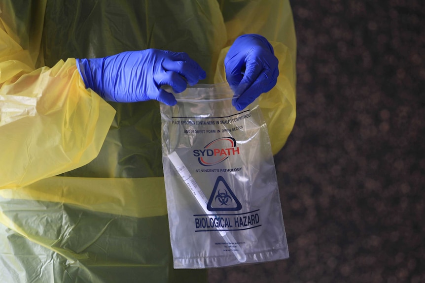 Health worker wearing blue gloves and yellow gown holds plastic sealed bag with swab test inside.