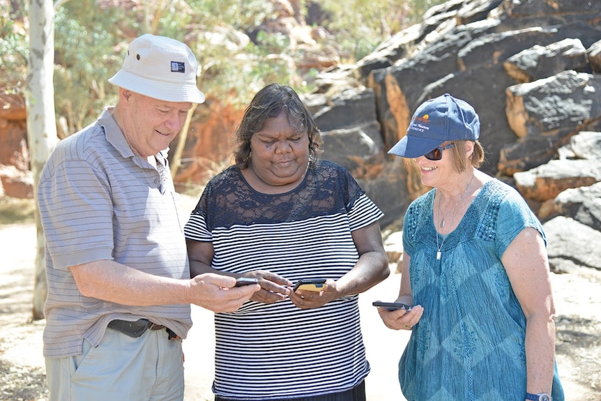 Two tourists stand on either side of traditional aboriginal owner Lynette Ellis who's showing them how to use an app on a phone.