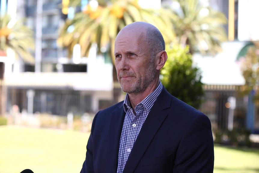 A bald man in a suit with no tie stands at a park.