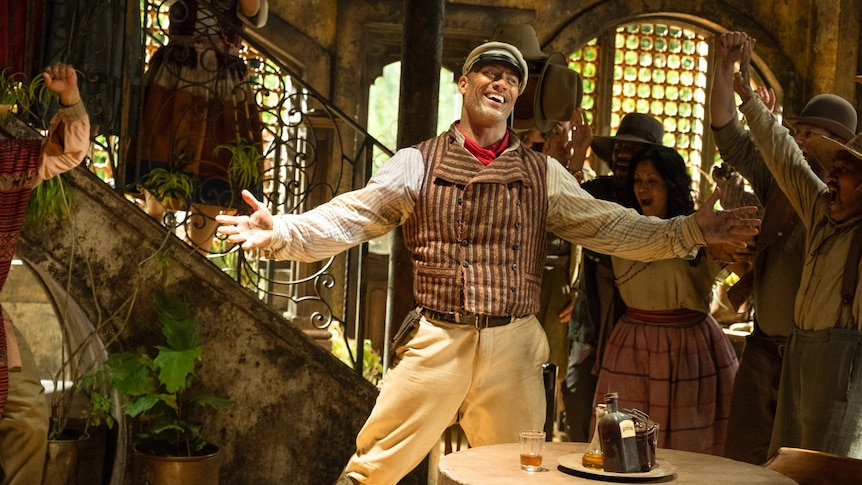 Dwayne Johnson smiles widely, his arms outstretched, standing inside an old-fashioned room with people celebrating behind him