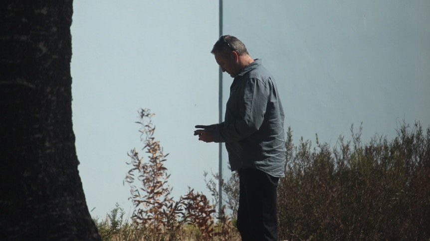 A middle-aged man stands near a tree, looking at his phone.