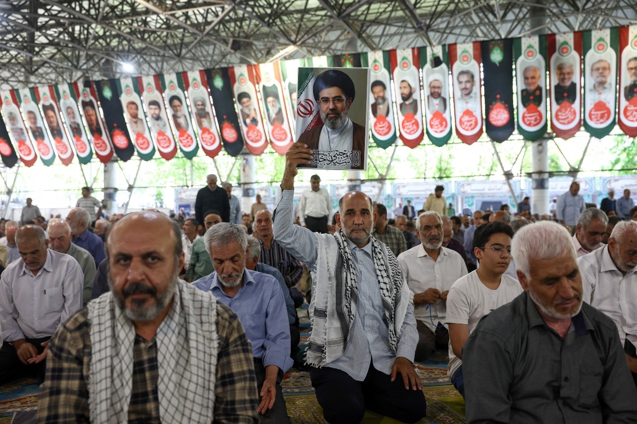 A large number of people praying. One is holding up a picture of the former leader of Iran
