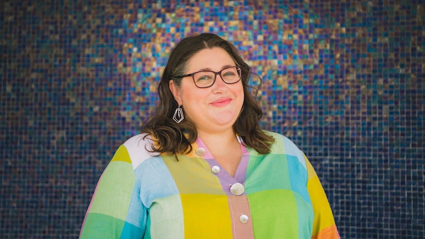 Katie wearing a colourful shirt and smiling in front of a mosaic-tiled wall.