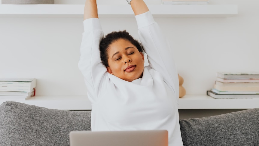 A woman stretching her arms, sitting in front of a computer in her home, for story about working from home rules.
