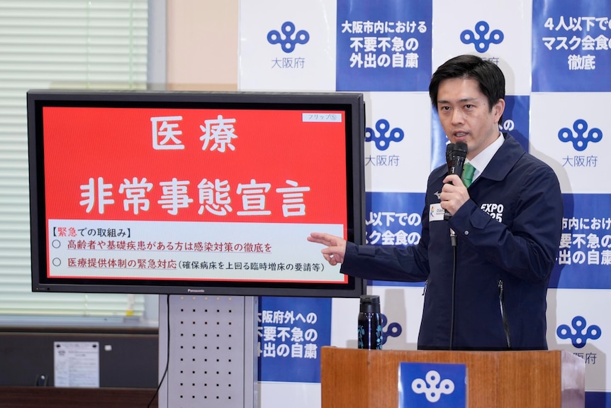 A governor speaks into a microphone next to a sign in red that says 'medical emergency' in Japanese.