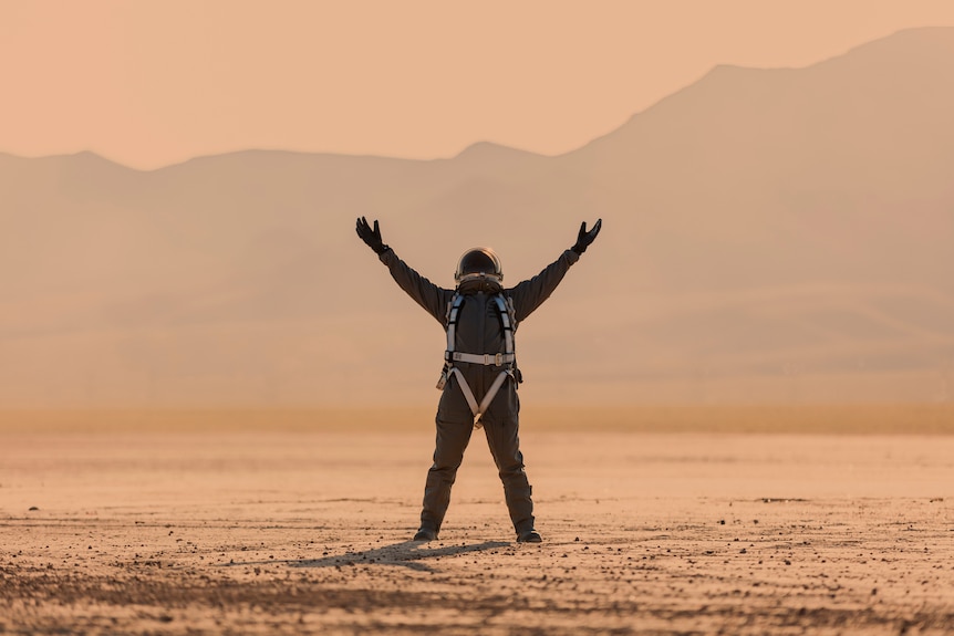 Profile of a person in a space suit looking upwards and arms outstretched to the sky. Mountainous Mars' landscape as backdrop.