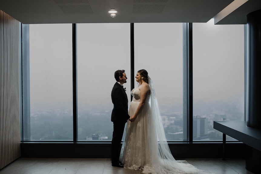 A man wearing a black suit and a woman in a white wedding dress stand in front of a large window facing each other