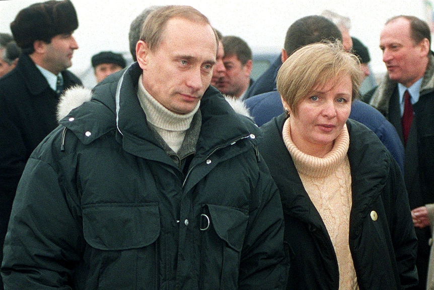 Vladimir Putin in an enormous navy puffer jacket and cream turtleneck standing next to a woman with cropped blonde hair