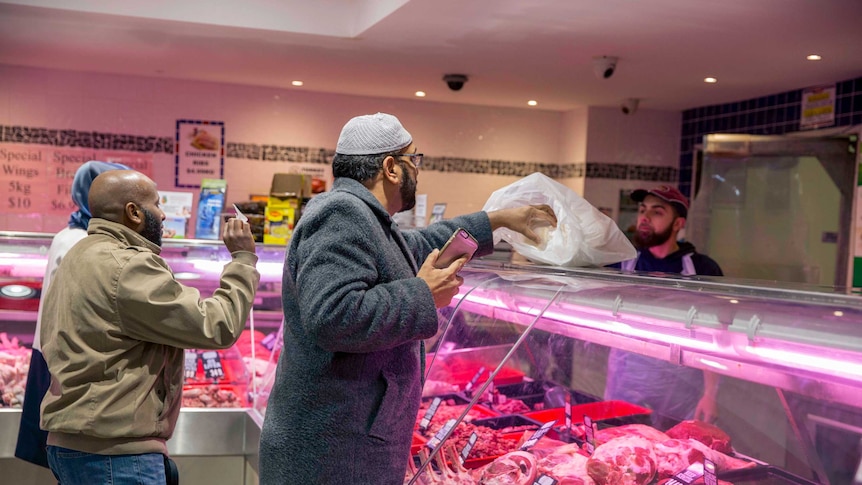 A man in a kufi and robes buys meat from a butcher.
