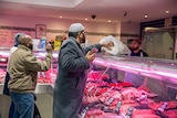 A man in a kufi and robes buys meat from a butcher.