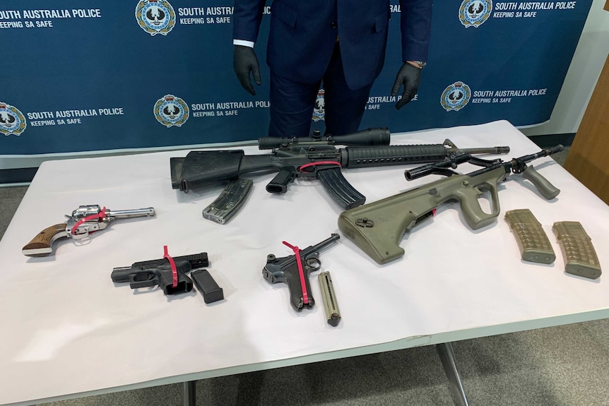 Two assault rifles and three handguns lay on a table with a police offi