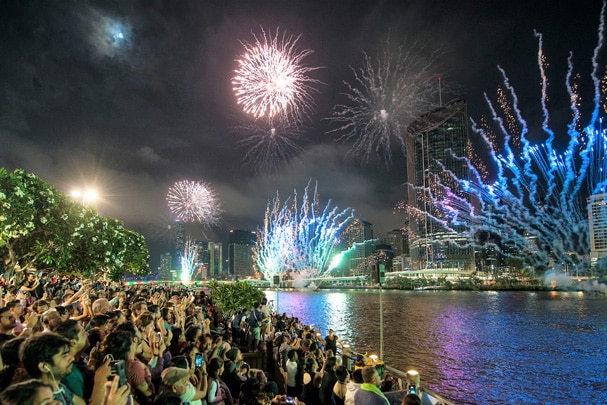 Hundreds of people standing in front of the river watch fireworks
