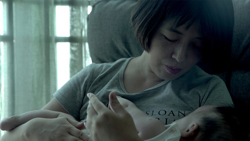 Colour close-up still of Gai Qi seated and cradling her baby near a window in 2019 documentary Leftover Women.