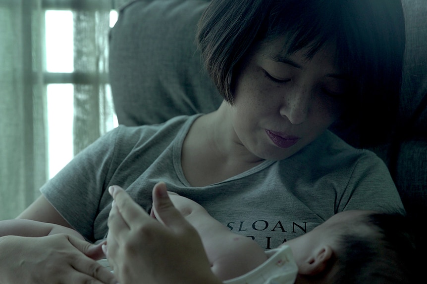 Colour close-up still of Gai Qi seated and cradling her baby near a window in 2019 documentary Leftover Women.