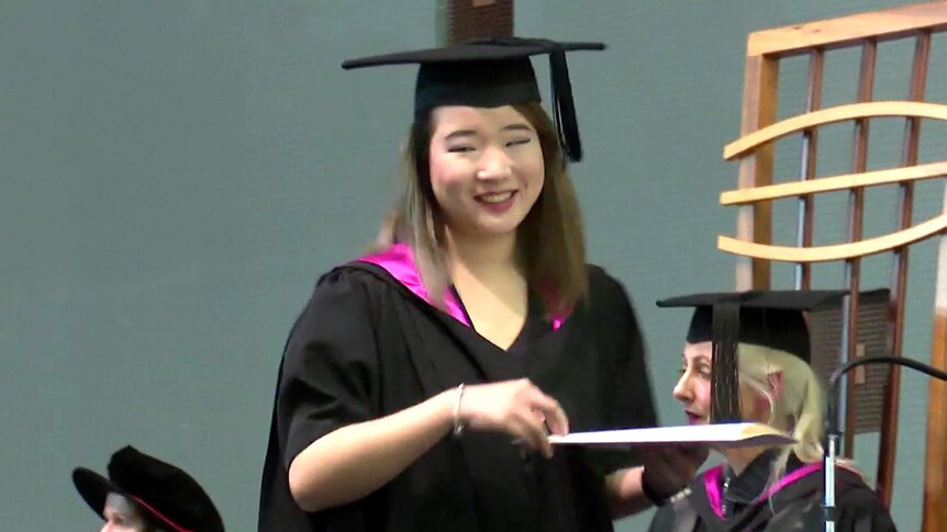 A young woman accepts a degree on stage while wearing graduation robes and a mortar board hat.