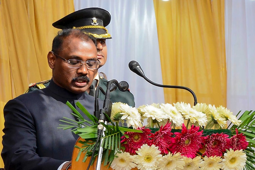 A man in a dark suit stands behind a lectern with bright flowers and speaks into a microphone as a soldier stands guard behind.