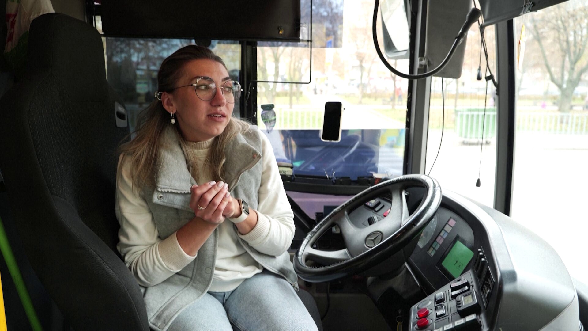 Woman sitting in the driver's seat of a bus talks, holding her hands. 