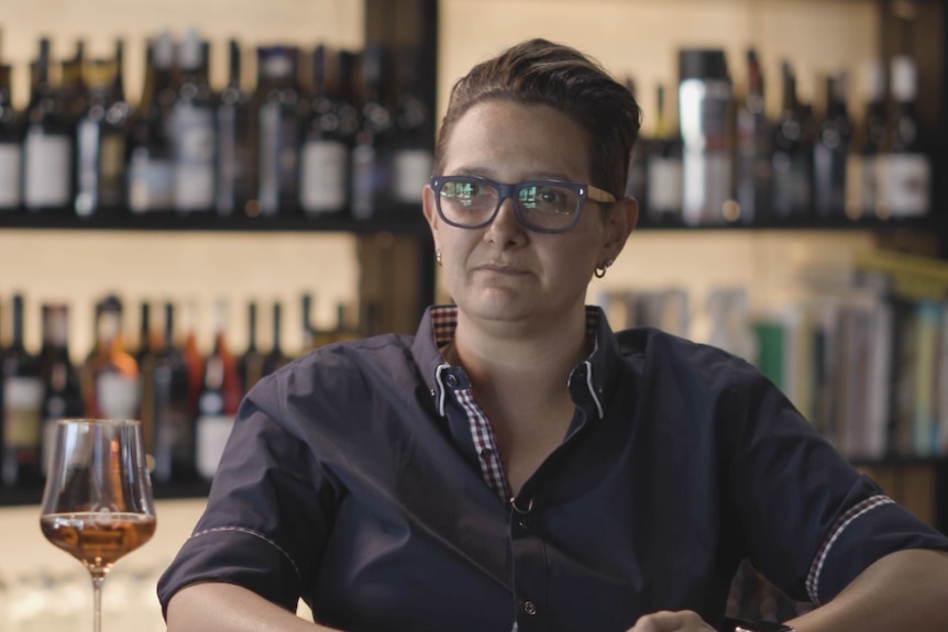 A woman in a blue shirt sits at a table with a glass of wine.