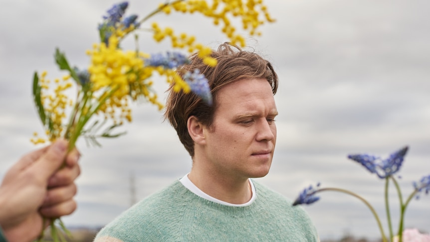 Tourist has blonde-brown short hair and wears a mint green jumper. He is pictured with yellow bottle brush and lavendar.