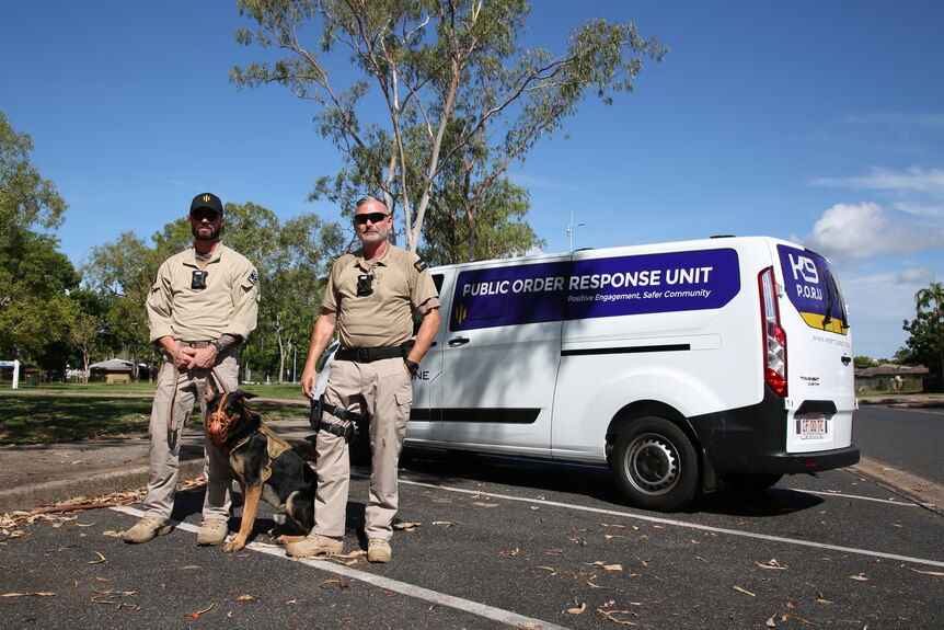 Two men wearing security uniforms with a muzzled dog, standing next to a van with 'Public Order Response Unit' on the side