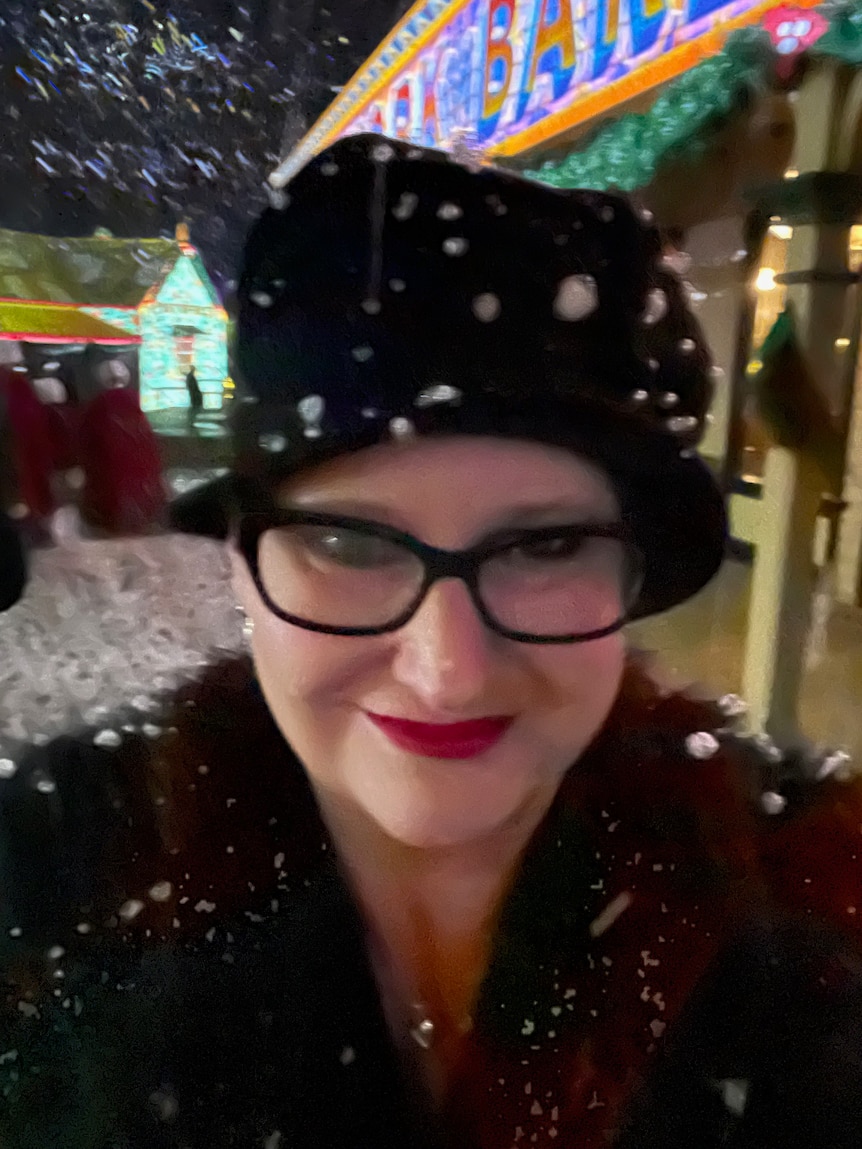A selfie of a smiling Caucasian woman in snow, night, wears black hat, red lipstick, fur collar, lit up sign behind.