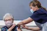 An older woman with grey hair, wearing a floral face mask and T-shirt, lifts a sleeve as a nurse injects the COVID vaccine