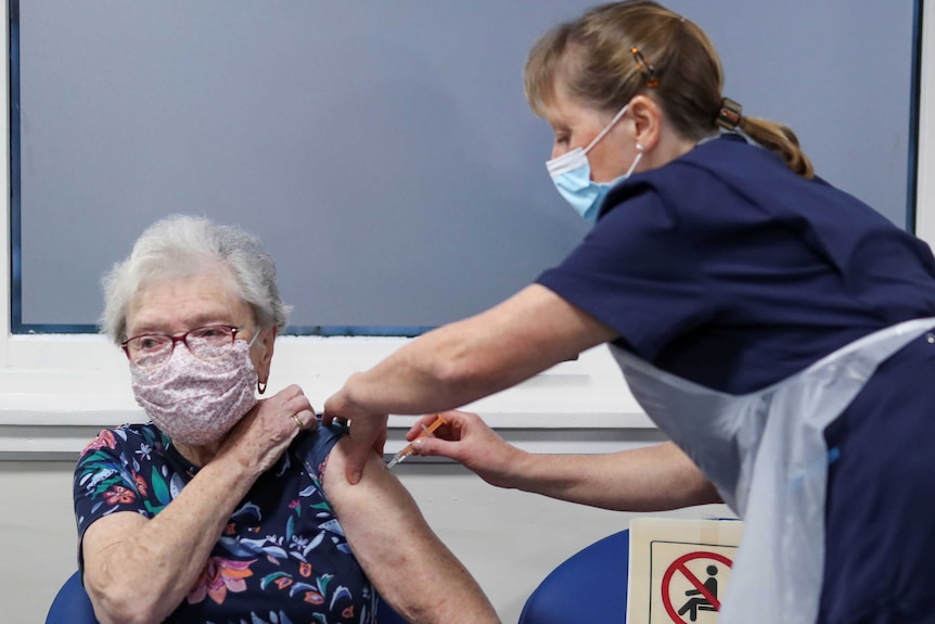 An older woman with grey hair, wearing a floral face mask and T-shirt, lifts a sleeve as a nurse injects the COVID vaccine