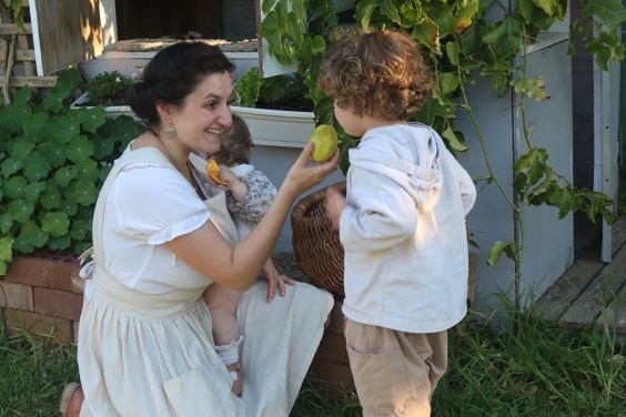 Outside in a bushy yard, a woman crouches down smiling, holding a baby, and holds an apple out towards another child. 
