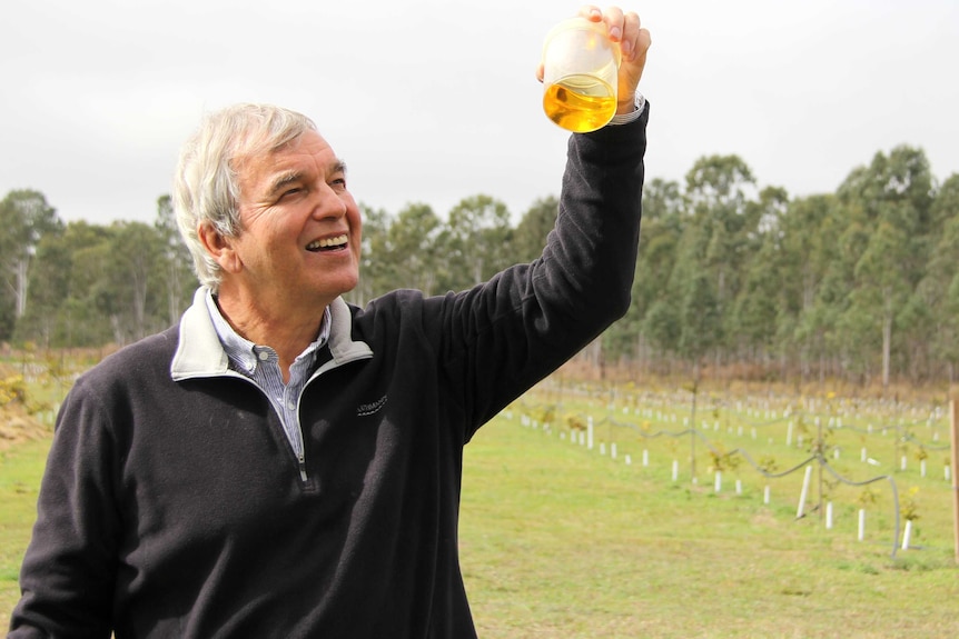 A smiling man with short, grey hair stands in a field and holds up a beaker of oil.